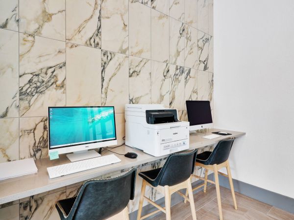 Two desktop computers and a printer on a desk with black chairs against a marble-tiled wall.