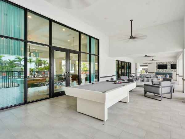 Modern indoor lounge with a white pool table, seating area, and large glass doors overlooking a pool.